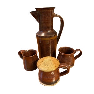 Puede incluir: Un juego de cuatro tazas de cerámica marrón con una jarra marrón y amarilla. Las tazas tienen una forma redondeada y un asa en el lateral. La jarra tiene un pico y un asa. Las tazas y la jarra tienen un esmalte moteado.