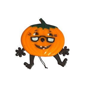 May include: A bright orange pumpkin ornament with a dark green stem, black limbs, and black hands. The pumpkin features a printed face with black-rimmed glasses, a smiling mouth with visible teeth, and a thin black string attached for hanging.