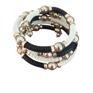 Puede incluir: Un conjunto de pulseras en espiral en negro, blanco y plateado. Las pulseras presentan una textura tejida con cuentas y detalles plateados. El diseño es moderno y adecuado para el uso diario.