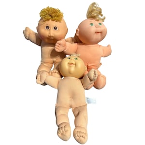May include: Three soft-bodied dolls with vinyl heads and yarn or moulded hair. One doll has curly golden yarn hair and brown eyes, another has blonde hair and green eyes, and the third has short blonde hair and green eyes. All have peach skin.