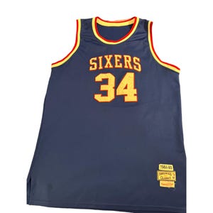Puede incluir: Una camiseta de baloncesto azul marino con la palabra "SIXERS" y el número "34" en amarillo. La camiseta tiene ribetes amarillos y rojos alrededor del cuello y las sisas. Una etiqueta "1984-85 Hardwood Classics Mitchell & Ness" está en la parte inferior.