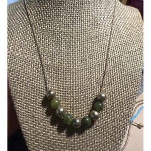 Puede incluir: Un collar con una fina cadena dorada. El collar presenta una serie de cuentas redondas de color verde oliva intercaladas con cuentas más grandes de color dorado metálico. El collar se exhibe sobre una superficie con textura de arpillera.