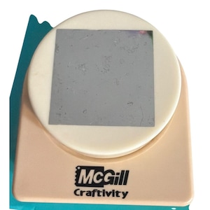 May include: A beige and cream-colored McGill Craftivity paper punch featuring a large, circular top button with a square silver-colored metal plate in the center. The brand name McGill and the word Craftivity are printed in black on the front.