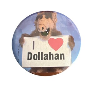 May include: A brown and gray alien character holding a sign that says "I ♥ Dollahan". The sign is white with a red heart.