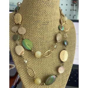 Puede incluir: Un collar dorado con una variedad de cuentas y colgantes. El collar presenta cuentas ovaladas, redondas y en forma de disco en tonos verdes, beige e iridiscentes. El collar se exhibe sobre un busto cubierto de arpillera.