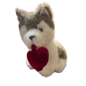 May include: A plush brown and white husky dog holding a red heart-shaped pillow. The dog is sitting and looking to the side.