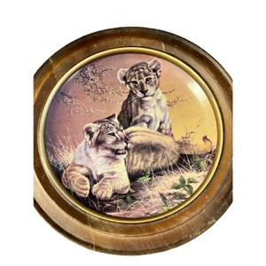 May include: A framed ceramic plate with a painting of two lion cubs in a grassy field. The cubs are looking at the viewer, and the background is a soft orange and yellow.