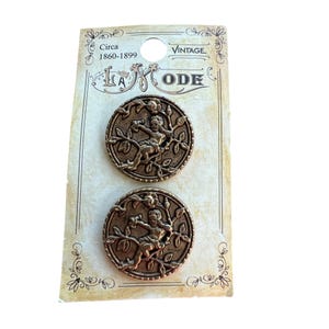 May include: Two antique gold-toned metal buttons featuring a raised design of a cherub sitting in a tree with leaves and flowers. The buttons are on a card that reads "Vintage Circa 1860-1899 La Mode".