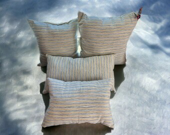 May include: Four striped throw pillows in shades of beige and blue. The pillows are stacked on top of each other, with the largest pillow at the bottom and the smallest pillow at the top.
