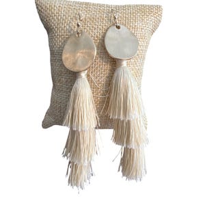 May include: A pair of gold-toned earrings with a layered tassel design. Each earring features a hammered, irregular-shaped gold disc attached to three tiers of cream-colored tassels. The earrings are displayed on a beige, textured surface.