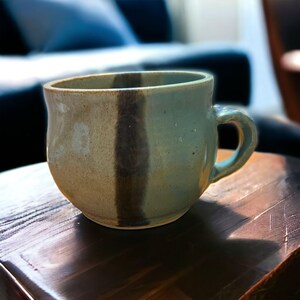Könnte beinhalten: Eine handgefertigte Keramik-Tasse mit rundem Korpus und einem stabilen Henkel. Die Tasse hat ein zweifarbiges Design mit einem vertikalen dunkelbraunen Streifen auf hellgrünem und beigem Hintergrund. Die Tasse steht auf einer Holzoberfläche.