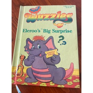 Könnte beinhalten: Ein Kinderbuchcover mit einem lila Elefanten mit rosa Streifen, der ein Los hält. Der Buchtitel lautet "Wuggles" und der Untertitel lautet "Eleroo's Big Surprise".