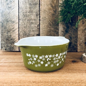 May include: A green casserole dish with a white floral pattern. The dish has a white interior and a rounded shape.