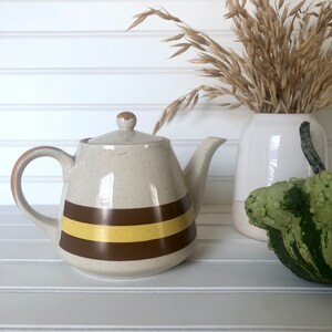 Puede incluir: Una tetera de color beige con manchas marrones y dos rayas amarillas y marrones alrededor del medio. La tetera tiene una tapa marrón y un asa marrón.