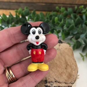 May include: A small ceramic figurine of Mickey Mouse. He is standing with his hands on his hips and wearing his signature red shorts with yellow buttons, black shoes, and white gloves.