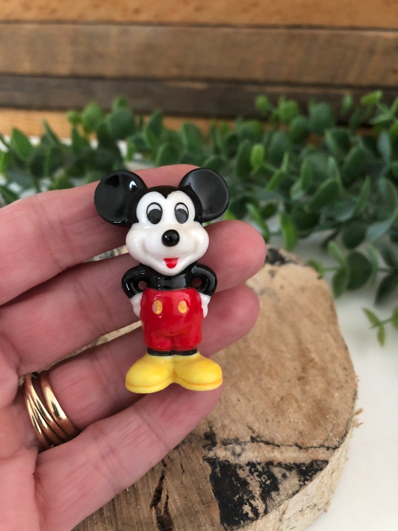May include: A small ceramic figurine of Mickey Mouse. He is standing with his hands on his hips and wearing his signature red shorts with yellow buttons, black shoes, and white gloves.