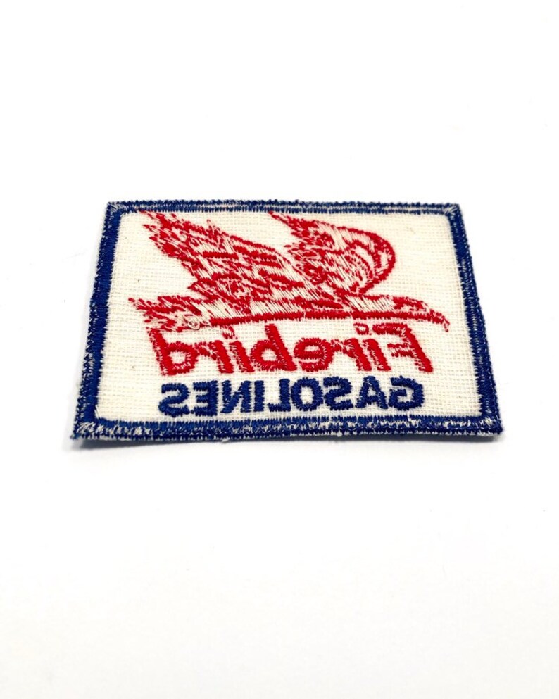May include: A white fabric patch with blue stitching around the edges. The patch features a red embroidered bird with the text "GASOLINE FIREBIRD" in red and blue embroidery.