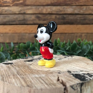 May include: A small black and white ceramic figurine of Mickey Mouse. He is wearing red shorts and yellow shoes. He is standing on a wooden surface.