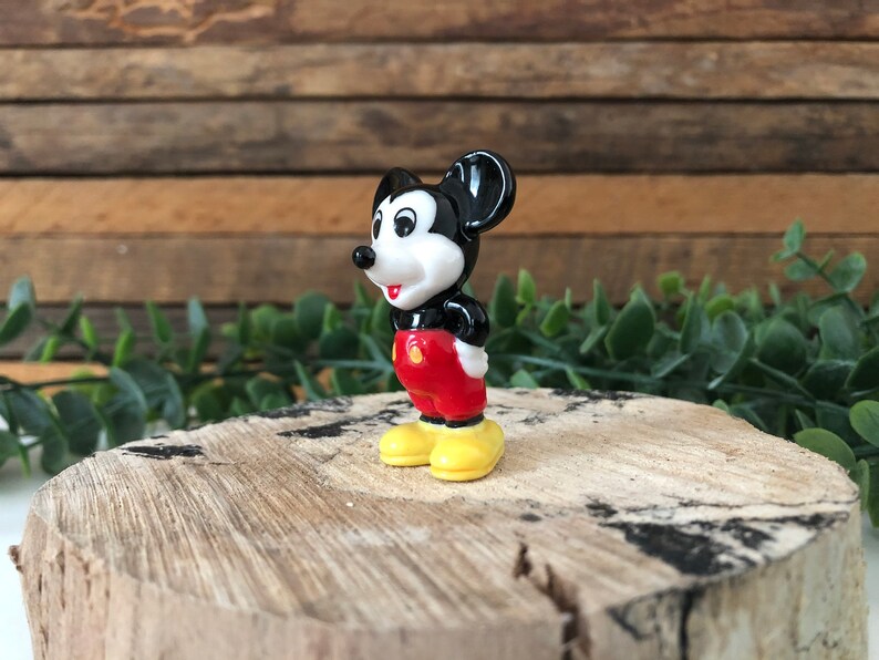 May include: A small black and white ceramic figurine of Mickey Mouse. He is wearing red shorts and yellow shoes. He is standing on a wooden surface.