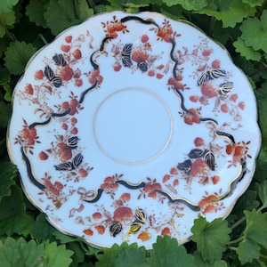 Puede incluir: Platillo de porcelana blanca con borde festoneado, adornado con un motivo floral en tonos naranja, marrón y dorado. Un diseño negro en forma de cinta con detalles dorados enmarca el arreglo floral. El platillo se encuentra sobre un fondo de follaje verde.