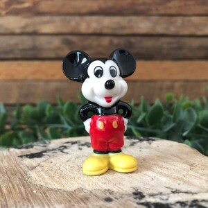 May include: A small, black and white ceramic figurine of Mickey Mouse. He is wearing red shorts with a yellow button and yellow shoes. He is standing with his hands on his hips.