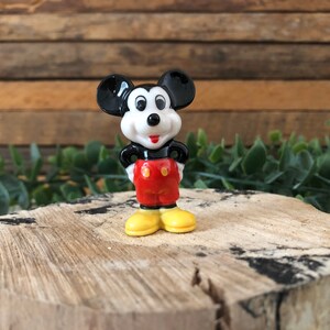 May include: A small ceramic figurine of Mickey Mouse. He is wearing his signature red shorts with yellow buttons and black shoes. He has a black and white face with big round ears.
