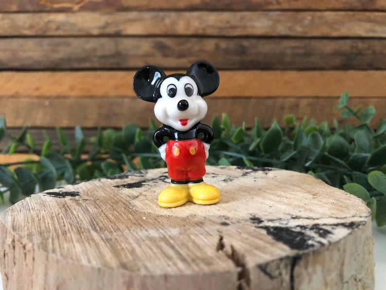 May include: A small ceramic figurine of Mickey Mouse. He is wearing his signature red shorts with yellow buttons and black shoes. He has a black and white face with big round ears.