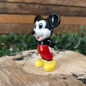 May include: A small ceramic figurine of Mickey Mouse. He is standing with his hands on his hips, wearing red shorts with yellow buttons and yellow shoes. He has black ears and a black nose.