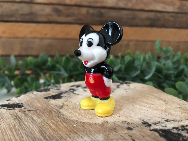 May include: A small ceramic figurine of Mickey Mouse. He is standing with his hands on his hips, wearing red shorts with yellow buttons and yellow shoes. He has black ears and a black nose.
