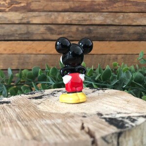 May include: A small ceramic figurine of Mickey Mouse. He is standing with his back to the camera, wearing his signature red shorts and yellow shoes. His black ears are visible.