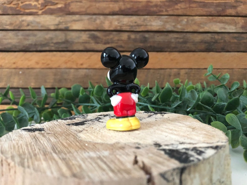 May include: A small ceramic figurine of Mickey Mouse. He is standing with his back to the camera, wearing his signature red shorts and yellow shoes. His black ears are visible.