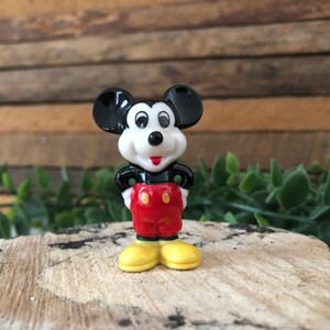 May include: A small ceramic figurine of Mickey Mouse. He is standing with his hands on his hips, wearing his signature red shorts with two yellow buttons and yellow shoes. His black ears are large and his face is white with black eyes and a red nose.