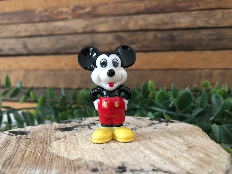 May include: A small ceramic figurine of Mickey Mouse. He is standing with his hands on his hips, wearing his signature red shorts with two yellow buttons and yellow shoes. His black ears are large and his face is white with black eyes and a red nose.