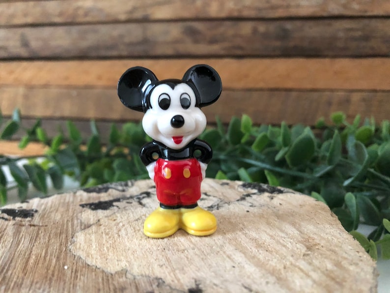 May include: A small, black and white ceramic figurine of Mickey Mouse. He is wearing red shorts with yellow buttons and yellow shoes. He is standing on a wooden surface.