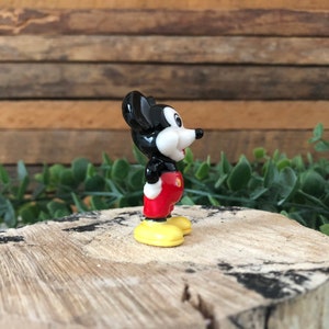 May include: A small ceramic figurine of Mickey Mouse. The figurine is black, white and red with yellow shoes. Mickey is standing with his right foot forward and his head turned to the right. He is smiling.