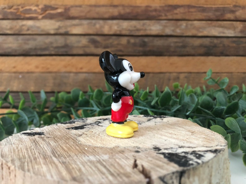 May include: A small ceramic figurine of Mickey Mouse. The figurine is black, white and red with yellow shoes. Mickey is standing with his right foot forward and his head turned to the right. He is smiling.