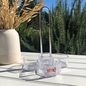 May include: A clear plastic condiment holder with a metal handle. The holder has three compartments and the word "HEINZ" is printed on the front.