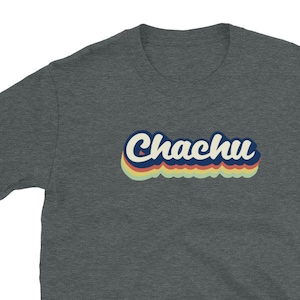 以下が含まれることがあります： レトロなフォントで「Chachu」という文字がプリントされたダークグレーのTシャツ。テキストは白で、青いアウトラインと多層の虹色の効果があります。このシャツは半袖で、柔らかい素材で作られています。