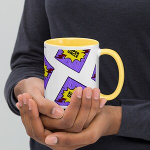 以下が含まれることがあります： 黄色い縁と取っ手のある白いセラミックマグカップ。マグカップには、「スーパーおじさん」と書かれた紫と黄色のコミックの吹き出しが繰り返しパターンで描かれています。