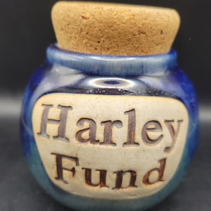 May include: A small, round, blue glazed ceramic jar with a cork stopper. The front features a recessed, unglazed beige oval panel with the embossed, dark-colored text "Harley Fund" centered on the surface of the jar.