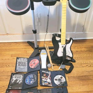May include: A video game drum kit with four colored pads, a black Fender Stratocaster guitar controller with a yellow neck, a foot pedal, two drumsticks, and a microphone. Four open game cases for Rock Band and Guitar Hero are displayed below.