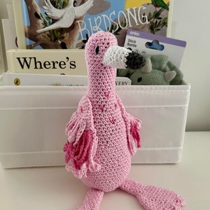 May include: A pink crocheted flamingo toy with a grey beak and pink and white wings. The toy is sitting on a white surface.
