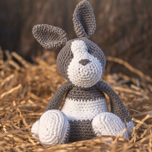 May include: A crocheted gray and white stuffed animal rabbit sitting in a bed of brown shredded paper.