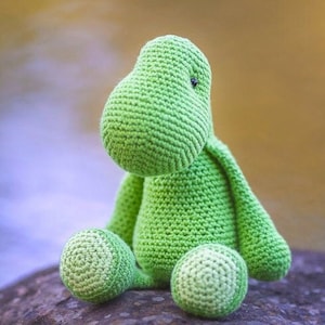 Puede incluir: Un juguete de dinosaurio de crochet verde sentado en una roca. El dinosaurio tiene una cabeza grande y brazos y piernas pequeños.