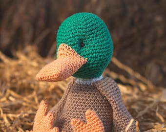Puede incluir: Un juguete de pato de crochet con cabeza verde, cuerpo marrón y pico y patas naranjas. El pato está sentado en un nido de papel triturado.