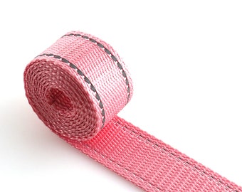 May include: Pink woven webbing with a black stitched trim. The webbing is rolled up and the end is laying flat.