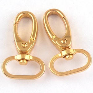 Pode incluir: Dois fechos giratórios de metal dourado com um anel em D em cada um. Os fechos estão abertos e prontos para serem presos a uma bolsa ou outro item.