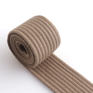 K&ouml;nnte beinhalten: Eine Rolle braunes Baumwollband mit gerippter Textur. Das Band ist aufgerollt und das Ende h&auml;ngt herunter.
