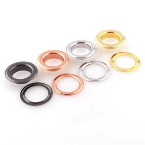 May include: Assortment of metal eyelets in various sizes and colors, including gold, silver, black, and copper. The eyelets are arranged on a white surface.