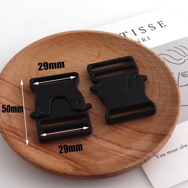 May include: Two black plastic side release buckles, each measuring 29mm wide and 50mm long. The buckles are on a wooden surface.